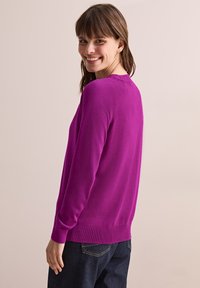 Pull vibrant en fuchsia, à manches longues, col rond, en tricot doux. Ourlet et poignets côtelés, texture lisse, porté sur un jean foncé.