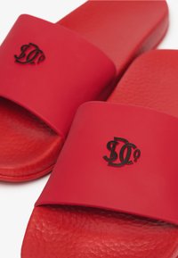 Mules rouges avec semelles texturées et larges brides présentant un logo "D" noir entrelacé sur chaque bride.