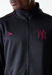 Giacca nera con zip e colletto alto, con logo dei New York Yankees in color bordeaux e strisce di accento laterali nello stesso colore. Tessuto liscio.