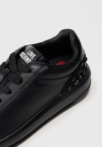 Sapatilha de couro preto com biqueira arredondada, atacadores na frente e sola de borracha. Apresenta a marca "LOVE MOSCHINO" e detalhes decorativos em preto no calcanhar.