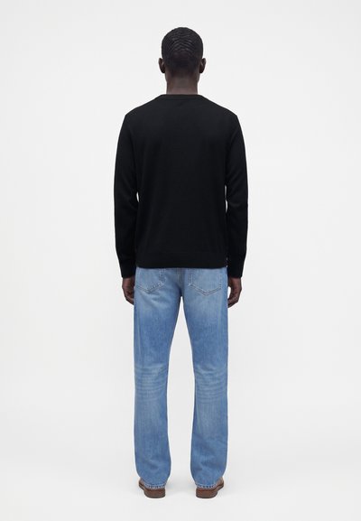Homme aux cheveux courts portant une chemise noire à manches longues, un jean bleu clair et des chaussures marron, debout de dos sur un fond blanc uni.