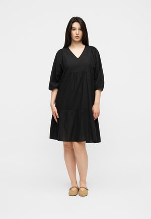 Femme aux longs cheveux foncés portant une robe noire à col en V longueur genou avec des manches bouffantes et des ballerines beiges tissées, debout sur un fond blanc.