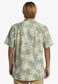 Persona con cabello castaño claro vistiendo una camisa de manga corta verde con un estampado tropical de hojas en crema y azul claro, vista desde atrás.