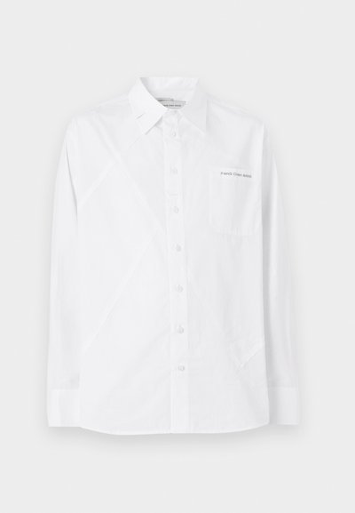 Camisa blanca de manga larga hecha de algodón, con cierre delantero de botones, un bolsillo en el pecho y detalles de costuras diagonales.