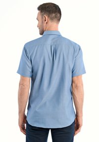 Lyhythihainen, vaaleansininen napapaita sileällä pinnalla, takasaumalla ja kaarevalla helmalla, miehen päällä, joka katsoo poispäin katsojasta.