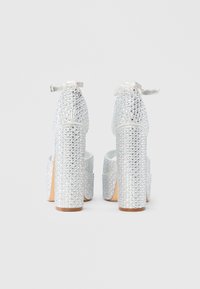 Srebrni čevlji z visokimi petami, okrašeni z bleščicami, s široko platformo in trakcem okoli gležnja z detajlom v obliki pentlje. Gladek tekstura in sijoč zaključek.