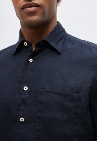 Άνδρας που φοράει σκούρα μπλε πουκάμισο με λευκά κουμπιά και τσέπη στο αριστερό στήθος, εμφανιζόμενος από τον λαιμό έως τη μέση.