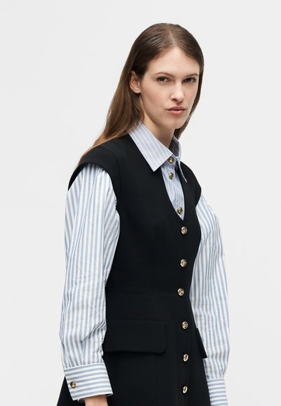 Femme aux longs cheveux bruns portant un gilet noir boutonné sur une chemise rayée bleue et blanche, sur un fond gris clair uni.