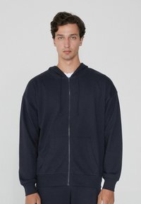 Sudadera de cremallera en azul marino, confeccionada en un tejido suave, con capucha, bolsillos delanteros y cordones ajustables. Presenta un corte relajado y puños acanalados.