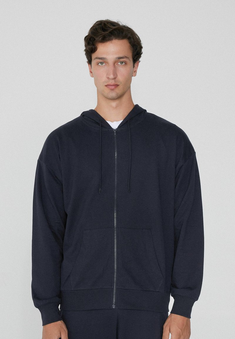 Sudadera de cremallera en azul marino, confeccionada en un tejido suave, con capucha, bolsillos delanteros y cordones ajustables. Presenta un corte relajado y puños acanalados.