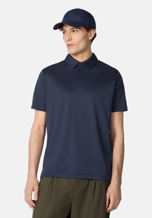 Uomo che indossa una polo blu navy e un cappellino abbinato, in piedi con le braccia rilassate, guardando leggermente di lato su uno sfondo semplice.