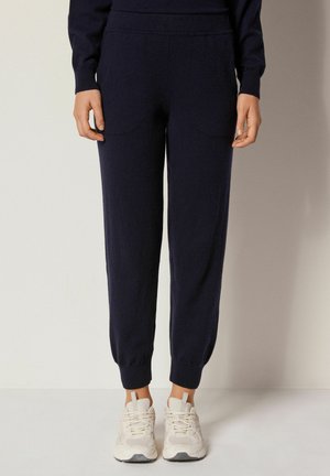 Pantaloni jogger a maglia blu navy, con una consistenza liscia, dotati di due tasche frontali e polsini elastici, abbinati a sneaker di colore chiaro.