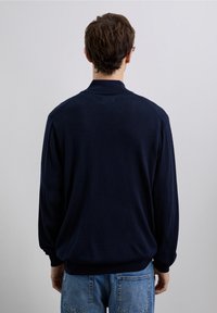 Granatowy sweter z dzianiny z prążkowanym kołnierzem i mankietami. Miękki materiał, luźny krój i minimalistyczne detale, zestawiony z jasnoniebieskimi dżinsami.