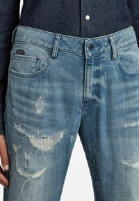 Light blue denim jeans with a distressed texture, featuring frayed patches and subtle fading. Five-pocket design with a button and zipper closure.