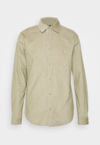 Camisa de pana en verde claro, con cuello estándar, mangas largas, cierre con botones y un pequeño parche de marca en el pecho.