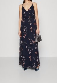 Blommig maxiklänning i marinblått med ljusrosa och vita blommor, har en djup V-ringning och tunna axelremmar, kombinerad med en liten grå handväska.