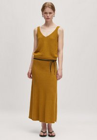 Mosterdgele gebreide tanktop en maxi-jurken set met een gestructureerde afwerking, gekenmerkt door een zwarte trekkoord aan de taille en platte zwarte sandaaltjes.