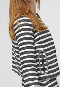 Paskiowana krótka kurtka w czerni i bieli, z teksturowanym splotem oraz zamykaną na zamek kieszenią z postrzępionymi krawędziami.