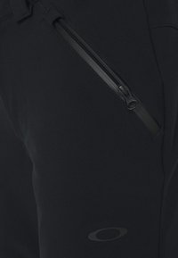 Gros plan sur un pantalon en tissu noir avec une poche zippée en diagonale et un petit logo ovale près du bas.