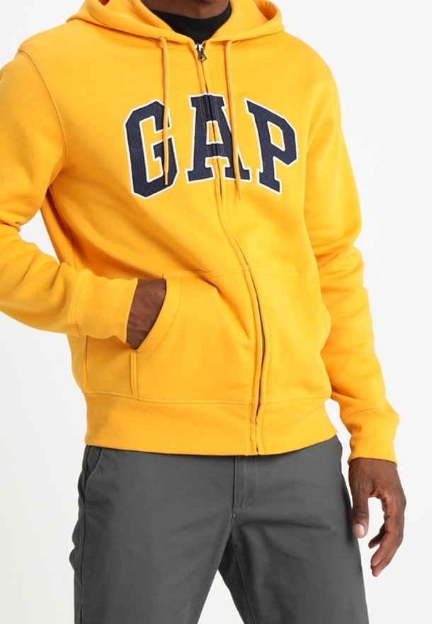 Sweat à capuche jaune avec fermeture éclair et grand logo "GAP" bleu marine. Il possède une poche kangourou et des poignets côtelés.