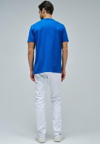 Camiseta azul de manga corta con escote redondo, combinada con unos jeans ajustados blancos y zapatillas blancas, visible desde la parte posterior.