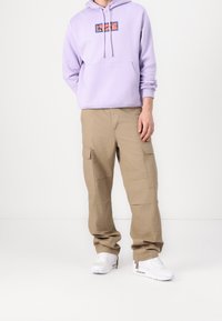 Ljuslila hoodie med grafisk tryck på bröstet, tillsammans med beige cargobyxor med flera fickor och vita sneakers.