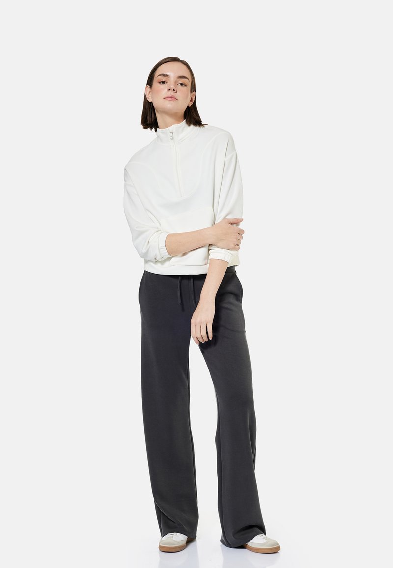 Woman standing with arms crossed wearing a white quarter-zip sweatshirt, black wide-leg pants, and white sneakers against a plain background.