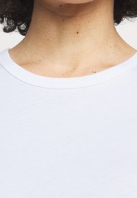 Vit bomull t-shirt med rund hals. Tyget verkar mjukt och slätt, med små texturdetaljer och prickar på huden nedan.