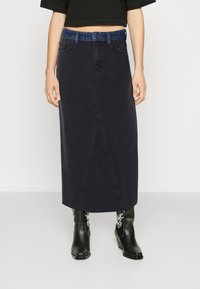 Donkerdenim maxi rok met een verborgen voornaad, een blauwe denim tailleband en een rechte snit. Draagt in combinatie met zwarte cowboystijl enkellaarzen.