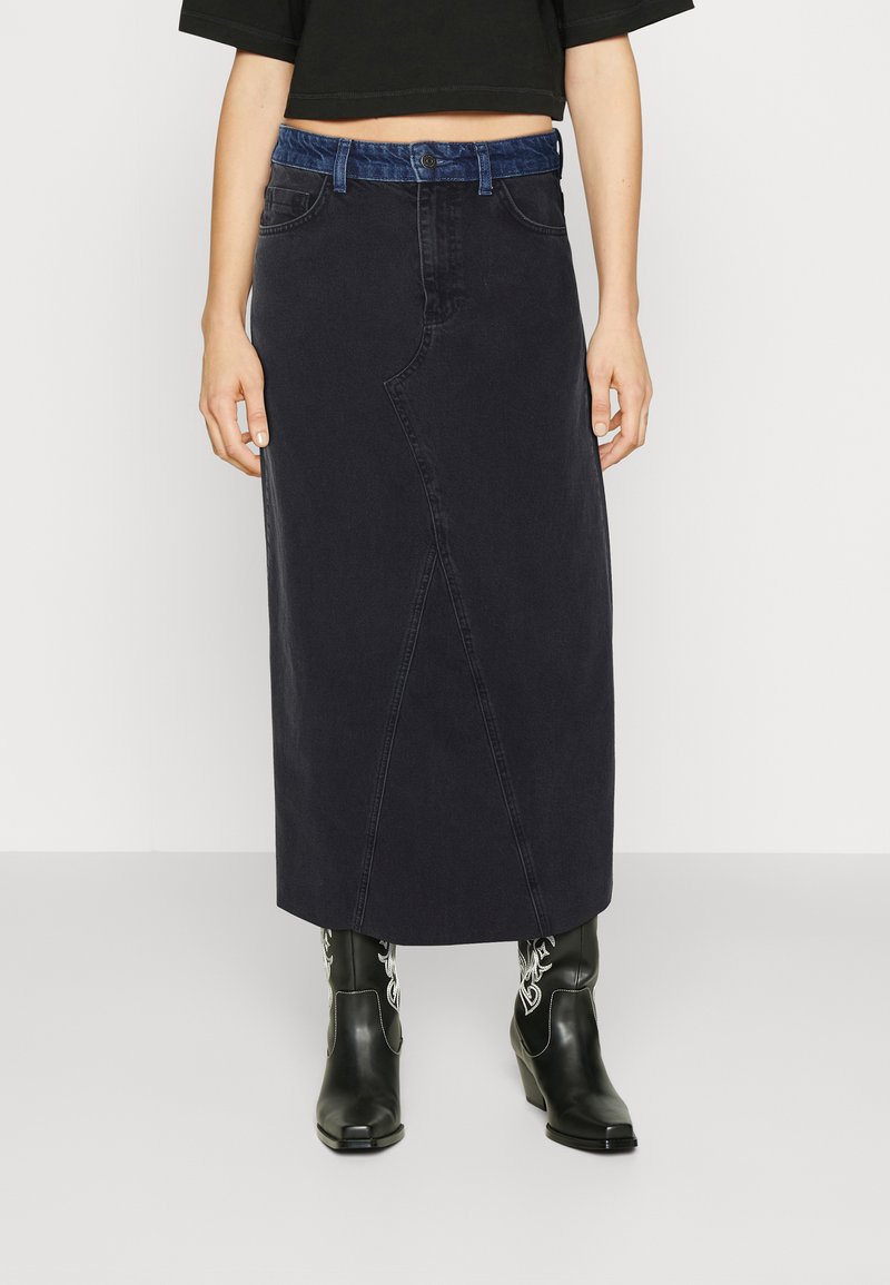 Donkerdenim maxi rok met een verborgen voornaad, een blauwe denim tailleband en een rechte snit. Draagt in combinatie met zwarte cowboystijl enkellaarzen.