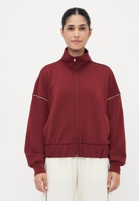 Bordeaux zip-up jakke med høy krage, ribbede mansjetter og kontrasterende piping langs skuldrene. Laget av mykt, lett stoff.