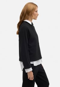 Top negro recortado con mangas largas y cuello redondo, superpuesto a una camisa blanca con cuello, llevado con pantalones negros. Textura suave.