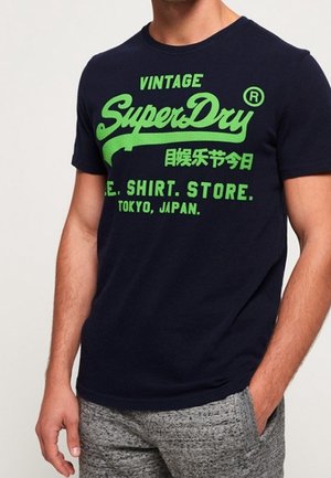 Mężczyzna w granatowej koszulce z jasną zieloną napisą „Vintage SuperDry Tokyo Japan” i szarych melanżowych spodniach, z ręką w kieszeni.