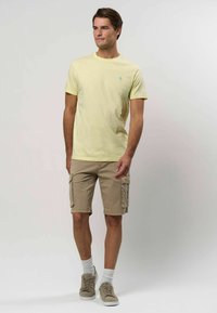 Heled kollane puuvillane t-särk, millel on väike türkiissinine logo, koos beeži värvi cargo-lühikeste pükste, millel on küljetaskud ja keeratud servad.