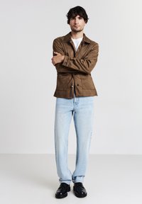 Giacca in suede marrone con colletto, due tasche frontali, abbinata a jeans azzurri chiari e scarpe nere. Design semplice e contemporaneo.