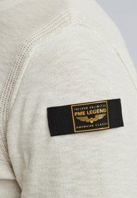 Tkanina boje krem s teksturiranim pletivom, s crnom oznakom na ramenu s natpisom "PME LEGEND" i zlatnim detaljima.