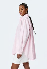 Oversized pink shirt made of lightweight fabric with white heart patterns, featuring a classic collar and button cuffs, with a curved hemline.