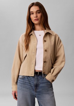 Frau mit langen braunen Haaren, die eine beige geknöpfte Jacke über einem weißen Crop-Top und blauen Jeans trägt, mit einer Hand in der Tasche.