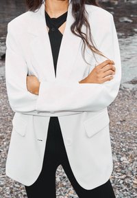 Biała marynarka o luźnym kroju, z wyłogami, bocznymi kieszeniami oraz zapięciem na jeden guzik, noszona na czarnej, fakturowanej bluzce i spodniach.