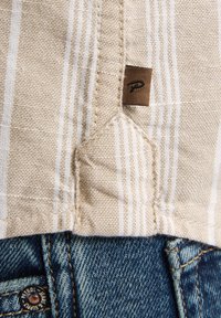 Tela de rayas beige con un tejido texturizado, que presenta una etiqueta marrón. Debajo, denim azul con costuras amarillas visibles, incluyendo un botón de marca.