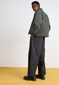 Olive green quilted bomber jacket with a ribbed collar, paired with loose-fitting dark trousers and black shoes on a yellow surface.