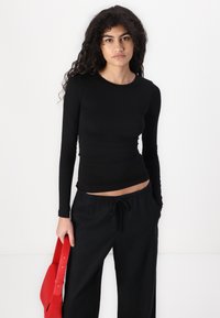 Crna majica dugih rukava u uskom kroju, izrađena od rastezljivog materijala. Uparena sa širokim crnim hlačama i crvenom torbom.