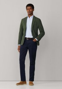 Mann trägt einen grünen Blazer, hellblaues Hemd, marineblaue Hose, braunen geflochtenen Gürtel und beige Wildleder-Slipper und steht vor einem schlichten Hintergrund.
