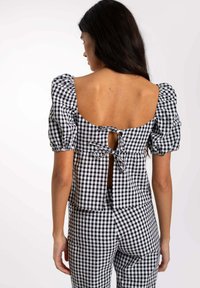 Blouse en vichy noir et blanc avec manches bouffantes et dos ouvert, dotée d'une fermeture à nœud. Associée à un bas vichy assorti.