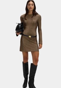 Bronzefarbenes langärmeliges Kleid mit taillierter Silhouette, schwarzem Gürtel mit runder Schnalle, schwarzer Handtasche und hohen schwarzen Stiefeln. Glatte, metallische Textur.