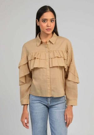 Femme aux longs cheveux bruns portant un chemisier beige à boutons avec des volants sur la poitrine et les manches, associé à un jean bleu clair.