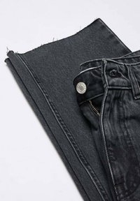 Vaqueros de mezclilla negra con herrajes plateados, con cierre de cremallera y botón, colocados junto a un trozo de tela a juego.