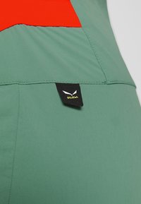 Gros plan sur un tissu vert et rouge avec une étiquette noire de la marque Salewa cousue près d'une couture.