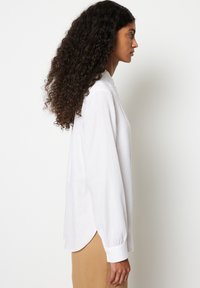 White long-sleeve shirt with a curved hem, soft texture, and buttoned cuffs, worn with beige pants, viewed from the side.