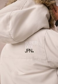 Beige jacket with a hood, featuring a faux fur trim. Embroidered "SUPER DRY" and "JPN" logos in white and black on the shoulder. Smooth texture.